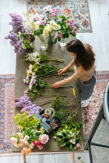 A woman arranging a variety of fresh flowers on a wooden table, including pink, white, and purple blossoms such as roses, lisianthus, and lilacs, with green foliage. The flowers are organized in small